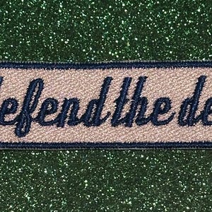 May include: A rectangular patch with the words "defend the den" embroidered in navy blue script. The patch has a light beige background with a navy blue border. The background is a sparkly, glittery green.