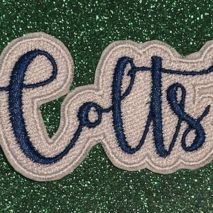 May include: Embroidered patch featuring the word "Colts" in a cursive font. The text is navy blue with a light gray outline and a white border. The patch is set against a sparkly green background.