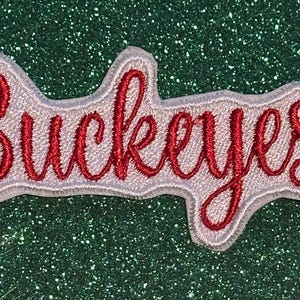 May include: Embroidered patch with the word "Buckeyes" in red script lettering, outlined in white. The patch is on a green glitter background. Ideal for jackets, bags, or other fabric items.