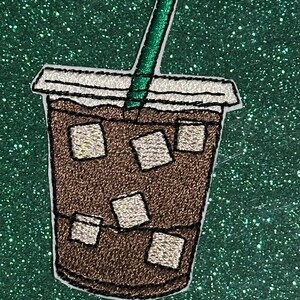May include: Embroidered patch of an iced coffee drink in a clear cup with a green straw. The coffee is brown with ice cubes. The cup has a white lid and is outlined in black. The background is a glittery green.