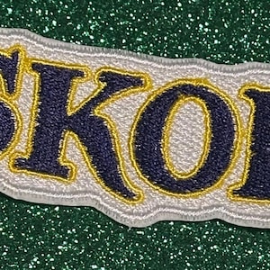 May include: Embroidered patch with the word "SKOL" in navy blue, outlined in gold, on a white and gray background. The patch has a wavy edge and is set against a glittery green surface.