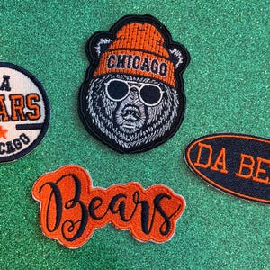 May include: Four embroidered patches featuring Chicago football themes. One circular patch reads DA BEARS CHICAGO with a star. One bear head patch wears an orange CHICAGO beanie and sunglasses. One oval patch reads DA BEARS. One orange script patch reads Bears.