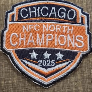 May include: Embroidered patch with a shield design. The patch features the word "CHICAGO" at the top, "NFC NORTH CHAMPIONS" in the center, and the year "2025" at the bottom. The patch has orange, navy blue, white, and black colors.
