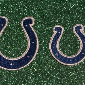 May include: Two navy blue horseshoe patches with silver trim, one large and one small, are displayed against a sparkly green background. The patches have a textured appearance and are likely for decorative purposes.