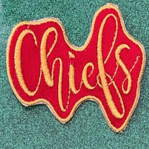 May include: A red embroidered patch with the word "Chiefs" in gold script. The patch has a unique shape and a gold border, set against a glittery green background.