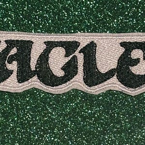 May include: Embroidered patch with the word "EAGLES" in black, outlined in a light gray color. The patch has a wavy border and is set against a green, glittery background.