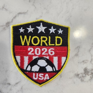 May include: Embroidered patch with a shield design. The patch features the text "WORLD 2026 USA" in yellow and white, with a soccer ball graphic. The shield is outlined in yellow, with a black background and red and white stripes.