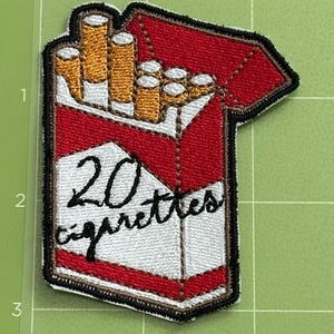 Pu&ograve; includere: Toppa ricamata raffigurante un pacchetto di sigarette. Il pacchetto &egrave; rosso e bianco, con la scritta "20 cigarettes" in nero. Le sigarette sono visibili, con filtri dorati. La toppa ha un bordo nero.