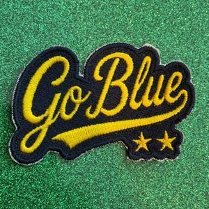May include: An embroidered patch featuring the text Go Blue in yellow cursive script on a dark navy blue background. The design includes a yellow underline extending from the text and two small yellow stars positioned below the lettering.