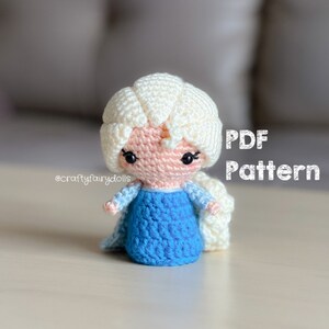Puede incluir: Mu&ntilde;eca amigurumi de crochet de un personaje animado popular con cabello largo y blanco, vestida con un vestido azul. La mu&ntilde;eca est&aacute; sentada sobre una superficie de madera clara. El texto "PDF Pattern" es visible en la imagen.