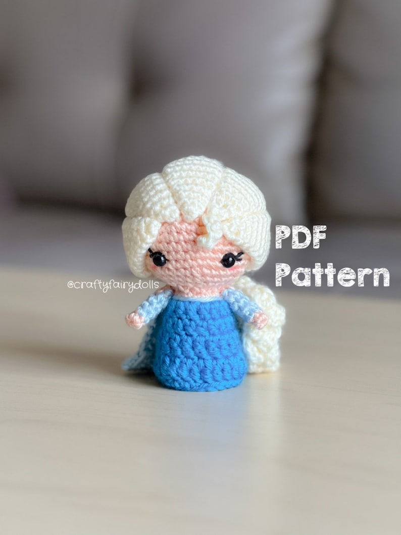 Puede incluir: Mu&ntilde;eca amigurumi de crochet de un personaje animado popular con cabello largo y blanco, vestida con un vestido azul. La mu&ntilde;eca est&aacute; sentada sobre una superficie de madera clara. El texto "PDF Pattern" es visible en la imagen.