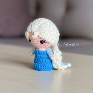 Puede incluir: Una mu&ntilde;eca de crochet con una trenza blanca, un vestido azul y una cara rosa. La mu&ntilde;eca tiene ojos negros y pesta&ntilde;as. La mu&ntilde;eca est&aacute; de pie sobre una superficie de madera. @craftyfairydolls