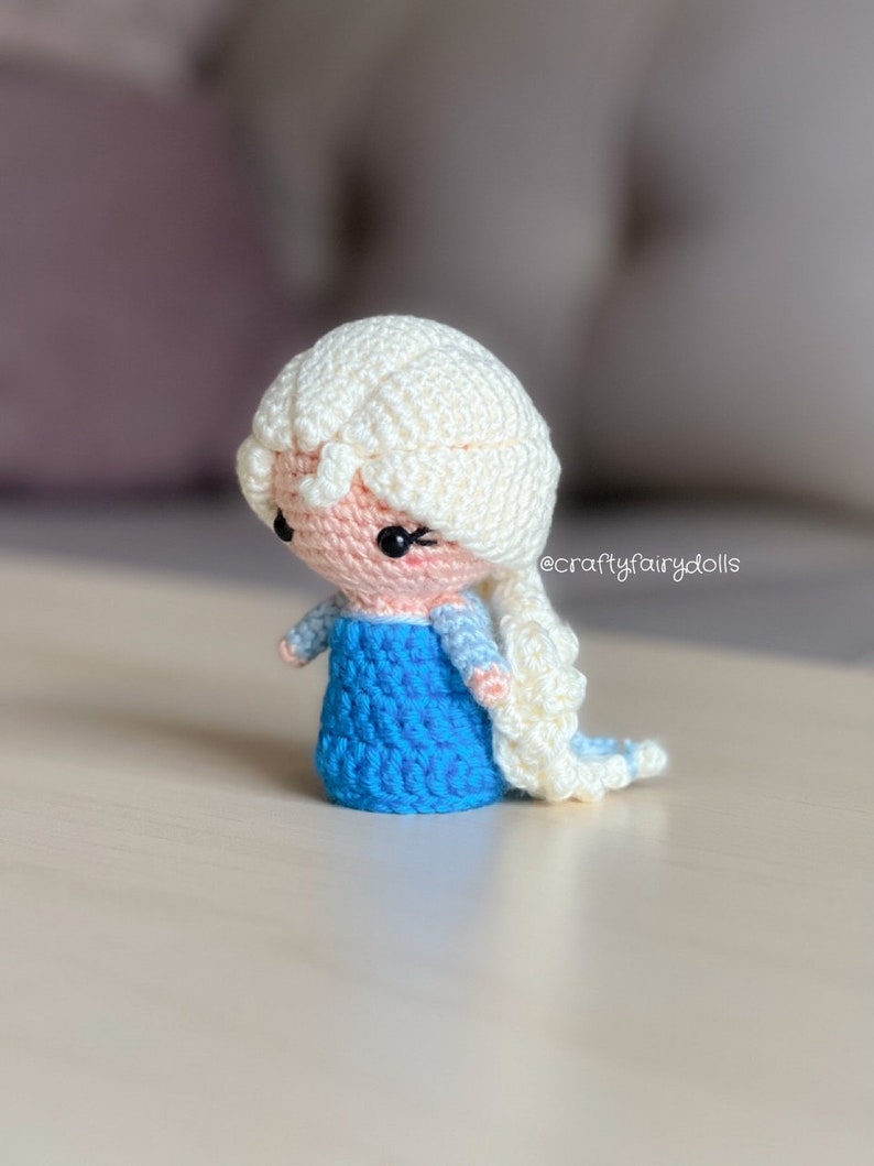 Puede incluir: Una mu&ntilde;eca de crochet con una trenza blanca, un vestido azul y una cara rosa. La mu&ntilde;eca tiene ojos negros y pesta&ntilde;as. La mu&ntilde;eca est&aacute; de pie sobre una superficie de madera. @craftyfairydolls