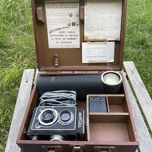 May include: Vintage Semflex camera in a brown case, open to reveal the camera, lens, and accessories. The case has a handle and metal clasps. The camera is black with silver accents. The case contains documents with French text.