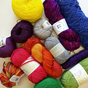 Op de afbeelding: Een verzameling kleurrijke garenbollen in verschillende tinten paars, geel, oranje, rood, groen en blauw. Het garen is gerangschikt op een wit oppervlak. Sommige garenbollen hebben labels met de woorden "Fiesta" en "Simply Recycled".