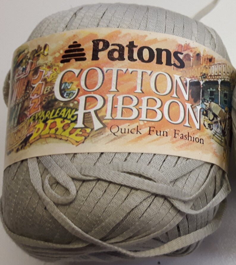 Op de afbeelding: Een bol Patons Cotton Ribbon garen in lichtgrijs. Het etiket luidt "Patons Cotton Ribbon Quick Fun Fashion".