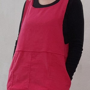 Op de afbeelding: Een vrouw draagt een roze corduroy schortjurk over een zwart longsleeve shirt. De jurk heeft twee zakken en een knoopdetail op de bandjes.