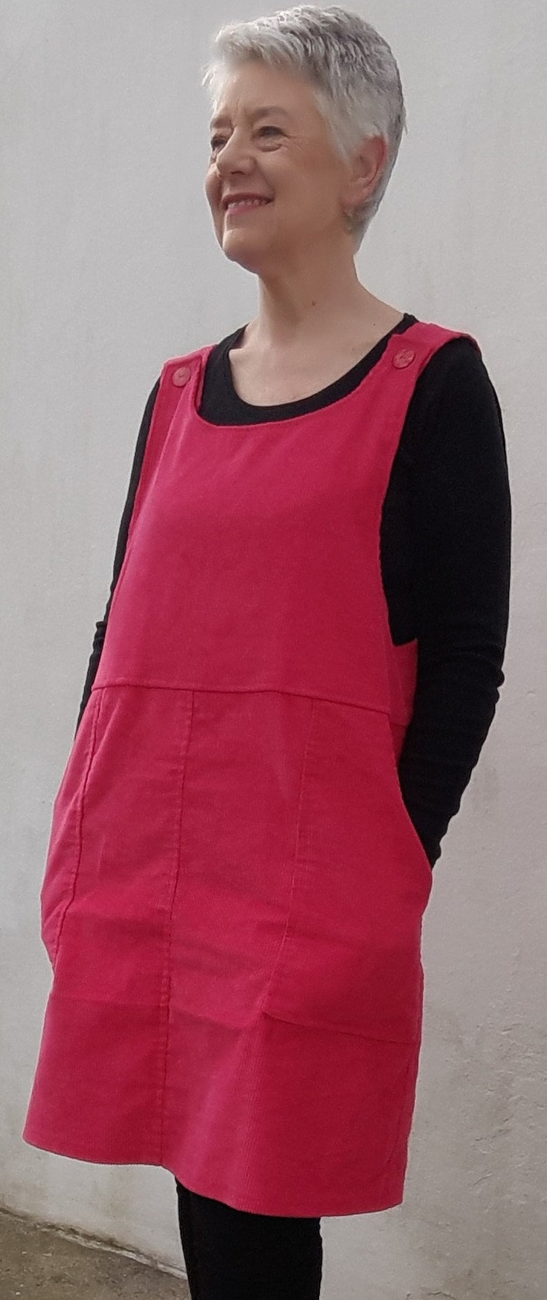 Op de afbeelding: Een vrouw draagt een roze corduroy schortjurk over een zwart longsleeve shirt. De jurk heeft twee zakken en een knoopdetail op de bandjes.