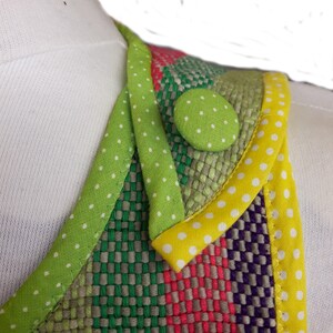 Op de afbeelding: Een close-up van een kleurrijke stof met een groene, gele en witte stippenbies. Een groene knoop met witte stippen is op de stof genaaid.