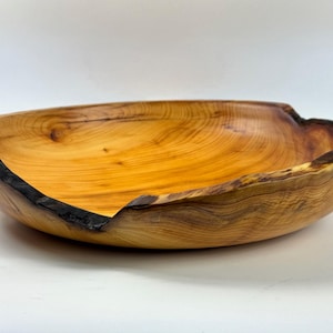 May include: A handcrafted wooden bowl with a natural edge. The bowl is made of light brown wood with a warm, golden hue and dark brown accents. The bowl's organic shape and unique grain patterns are visible.