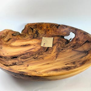 May include: A large, hand-carved wooden bowl with a natural, organic shape. The bowl features a rich, warm color palette of browns and tans, with intricate wood grain patterns. A small tag is affixed to the bowl. The bowl has a unique, irregular edge.