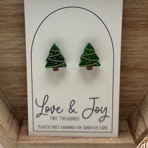 May include: A pair of Christmas tree earrings on a white card with the text "Love & Joy Tiny Treasures". The earrings are green with yellow and brown accents. The card is displayed in a wooden hexagonal tray.