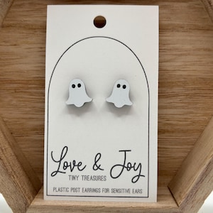 May include: A pair of white ghost-shaped stud earrings with black dot eyes on a white card. The card has the text "Love & Joy Tiny Treasures Plastic Post Earrings for Sensitive Ears". The earrings are displayed in a wooden hexagonal frame.