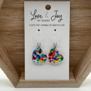 May include: A pair of heart-shaped earrings with a colorful confetti pattern. The earrings are made of clear acrylic and have silver posts. The card reads "Love & Joy Tiny Treasures Plastic Post Earrings for Sensitive Ears".