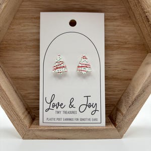 May include: White Christmas tree stud earrings with red and green accents. The earrings are displayed on a white card with the text "Love & Joy Tiny Treasures Plastic Post Earrings for Sensitive Ears". The earrings are set in a wooden display.