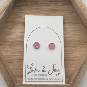 May include: A pair of pink stud earrings on a white card with the text "Love & Joy" and "Tiny Treasures". The card is inside a wooden hexagonal tray. The earrings are plastic post earrings for sensitive ears.