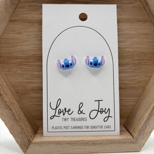 May include: A pair of blue and pink Stitch stud earrings on a white card. The card reads "Love & Joy Tiny Treasures Plastic Post Earrings for Sensitive Ears". The earrings are displayed on a wooden shelf.