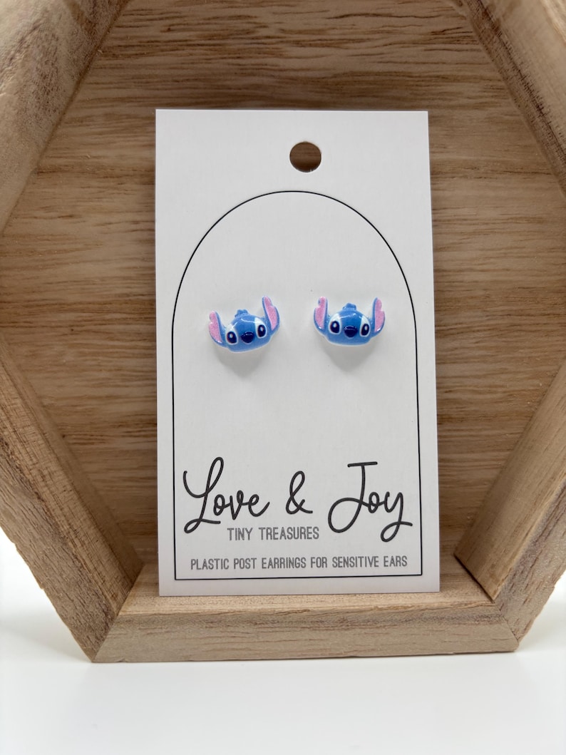 May include: A pair of blue and pink Stitch stud earrings on a white card. The card reads "Love & Joy Tiny Treasures Plastic Post Earrings for Sensitive Ears". The earrings are displayed on a wooden shelf.