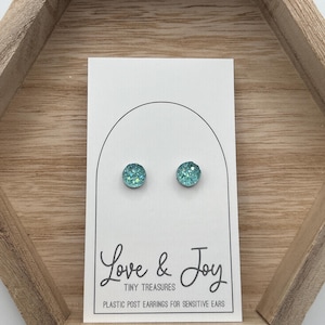 May include: A pair of teal-colored stud earrings with a glittery, textured surface. The earrings are displayed on a white card with the text "Love & Joy" and "Tiny Treasures". The card is inside a wooden hexagonal tray.