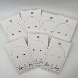 May include: A collection of stud earrings in various colors, including red, green, pink, blue, black, and clear. Each pair is displayed on a white card with the text "Love & Joy" and "Tiny Treasures".