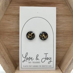 May include: A pair of round stud earrings featuring a black base with embedded gold flakes. The earrings are displayed on a white card with the text Love & Joy, TINY TREASURES, PLASTIC POST EARRINGS FOR SENSITIVE EARS printed in black ink.