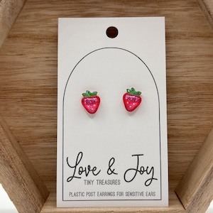 May include: A pair of red glitter strawberry stud earrings with green leaves. The earrings are on a white card with the text "Love & Joy Tiny Treasures Plastic Post Earrings for Sensitive Ears".