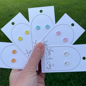 May include: Four pairs of smiley face stud earrings on white cards. The cards have the text "Love & Joy Tiny Treasures" printed on them. The earrings are in yellow, blue, pink, and white.
