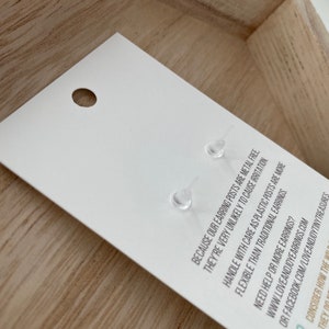 May include: A white earring card with two clear plastic earring posts. The card has text that reads, "BECAUSE OUR EARRING POSTS ARE METAL FREE, THEY'RE VERY UNLIKELY TO CAUSE IRRITATION." The card is in a wooden tray.