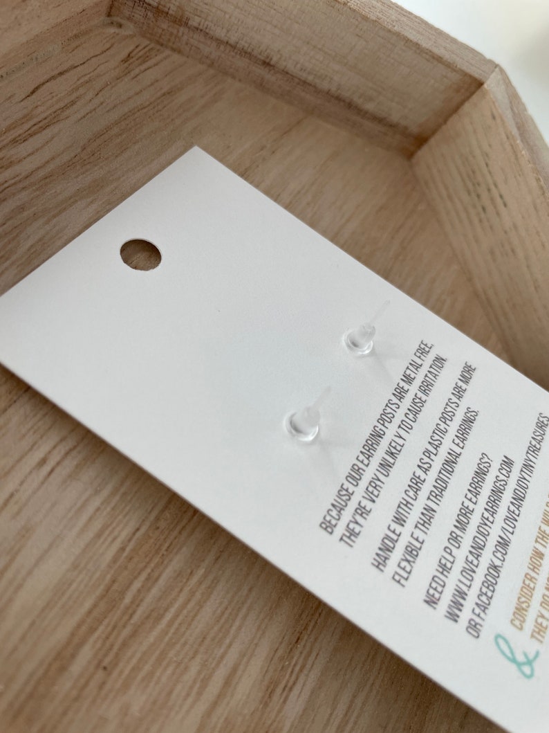May include: A white earring card with two clear plastic earring posts. The card has text that reads, "BECAUSE OUR EARRING POSTS ARE METAL FREE, THEY'RE VERY UNLIKELY TO CAUSE IRRITATION." The card is in a wooden tray.