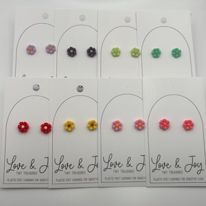 May include: Ten pairs of small flower stud earrings on white cards. The flowers are in various colors, including purple, green, yellow, pink, and red. The cards have the text "Love & Joy Tiny Treasures Plastic Post Earrings for Sensitive Ears".