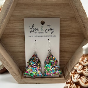 May include: A pair of Christmas tree-shaped earrings filled with colorful glitter. The earrings are clear with a silver hook and are displayed on a white card that reads "Love & Joy Tiny Treasures Plastic Post Earrings for Sensitive Ears."