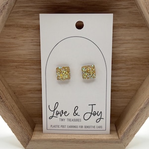 May include: A pair of square gold-toned druzy stud earrings on a white display card. The card features the text Love & Joy TINY TREASURES PLASTIC POST EARRINGS FOR SENSITIVE EARS. The earrings have a textured, sparkling surface finish.