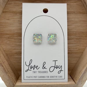 May include: A pair of square iridescent glitter earrings with plastic posts. The earrings are displayed on a white card with the text "Love & Joy Tiny Treasures Plastic Post Earrings for Sensitive Ears".