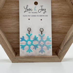 May include: Iridescent snowflake earrings on a white card with the text "Love & Joy Tiny Treasures Plastic Post Earrings for Sensitive Ears". The earrings have a blue and green color scheme and are displayed in a wooden tray.