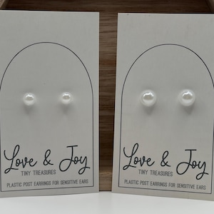 May include: Two pairs of white pearl stud earrings on white rectangular display cards. Each card features an arched line design and the text Love & Joy Tiny Treasures Plastic Post Earrings For Sensitive Ears printed in black ink.