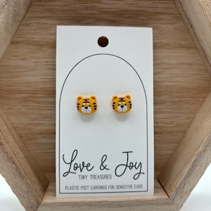 May include: A pair of yellow tiger face stud earrings with brown stripes and white accents. The earrings are on a white card with the text "Love & Joy Tiny Treasures Plastic Post Earrings for Sensitive Ears".