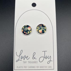 May include: A pair of small, round, floral print earrings with black backgrounds. The earrings are on a white card with the text "Love & Joy Tiny Treasures Plastic Post Earrings for Sensitive Ears".
