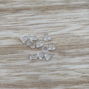 May include: A collection of clear, translucent earring backs scattered on a light brown wooden surface. The earring backs are small, with a rounded base and a post hole. They are made of a soft, flexible material.