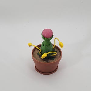 May include: A small, green, carnivorous plant with a pink, round, closed mouth and yellow tendrils in a brown pot.