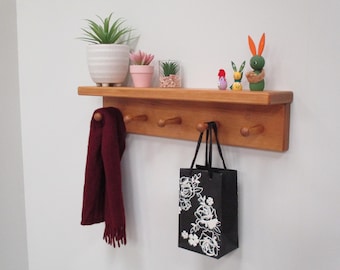 May include: Wooden wall shelf with four pegs, holding a burgundy scarf and a black gift bag with white floral design. The shelf displays potted plants and decorative wooden figurines. The shelf is mounted on a white wall.